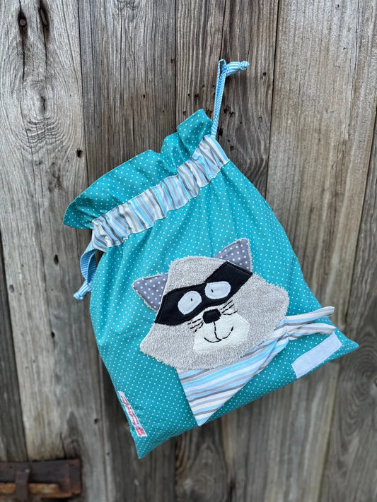 Türkiser Wetbag mit weißen Punkten, verziert mit einer bunten Stoffapplikation einem Waschbär grauem Frottee.DerBeutel hat einen gestreiften Tunnelzug und türkis karierte Kordeln, aufgenommen vor rustikalem Holzhintergrund.