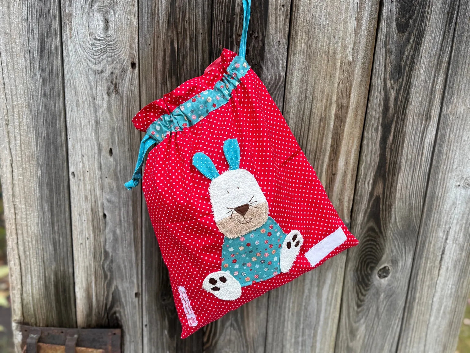 Roter Wetbag mit weißen Punkten, verziert mit einer bunten Stoffapplikation einem Hasen .DerBeutel hat einen floralen Tunnelzug und türkise Kordeln, aufgenommen vor rustikalem Holzhintergrund.