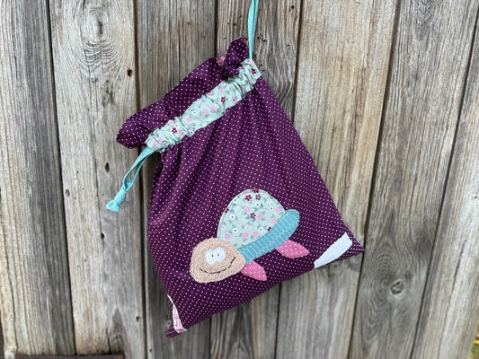 Dunkellila Wetbag mit weißen Punkten, verziert mit einer bunten Stoffapplikation einer Schildkröte aus floralen Stoff.DerBeutel hat einen floralen Tunnelzug und mintfarbene Kordeln, aufgenommen vor rustikalem Holzhintergrund.