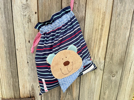 Dunkelblauer Wetbag mit weißen und roten Streifen, verziert mit einer bunten Stoffapplikation einem 
Bär.DerBeutel hat einen  Tunnelzug im graphischen Design und rot weiß karierte Kordeln, aufgenommen vor rustikalem Holzhintergrund.