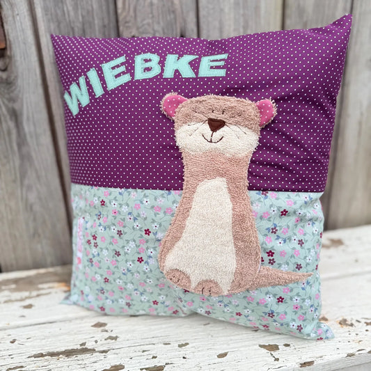 Handgenähtes Kinderkissen personalisiert, gestaltet dunkellila mit weißen Punkten im oberen Teil, altmint mit bunten Blümchen im unteren Teil. Auf dem Kissen ist eine Otter-Applikation in hellbraunem Frottee.Fotografiert vor einem rustikalen Holzhintergrund 