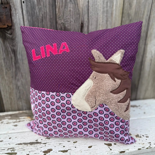Handgenähtes Kinderkissen personalisiert, gestaltet grün mit weißen Punkten im oberen Teil, lila mit pinken Blumen im unteren Teil. Auf dem Kissen ist eine Pferde-Applikation in hellbraunem Frottee.Fotografiert vor einem rustikalen Holzhintergrund 