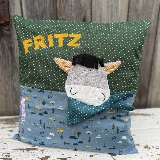 Handgenähtes Kinderkissen personalisiert, gestaltet grün mit weißen Punkten im oberen Teil, rauchblau mit Autoprint im unteren Teil. Auf dem Kissen ist eine Esel-Applikation in grauem Frottee.Fotografiert vor einem rustikalen Holzhintergrund 
