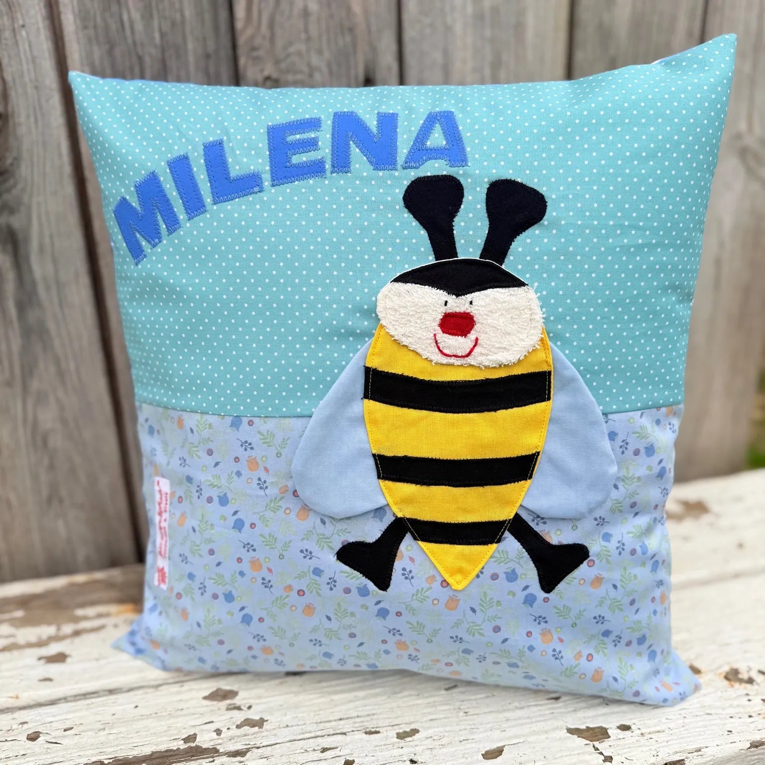 Handgenähtes Kinderkissen personalisiert, gestaltet mint mit weißen Punkten im oberen Teil, hellblau mit pastellfarbenen Blümchen bunten im unteren Teil. Auf dem Kissen ist eine Bienen-Applikation in leuchtendem Gelb. Fotografiert vor einem rustikalen Holzhintergrund 