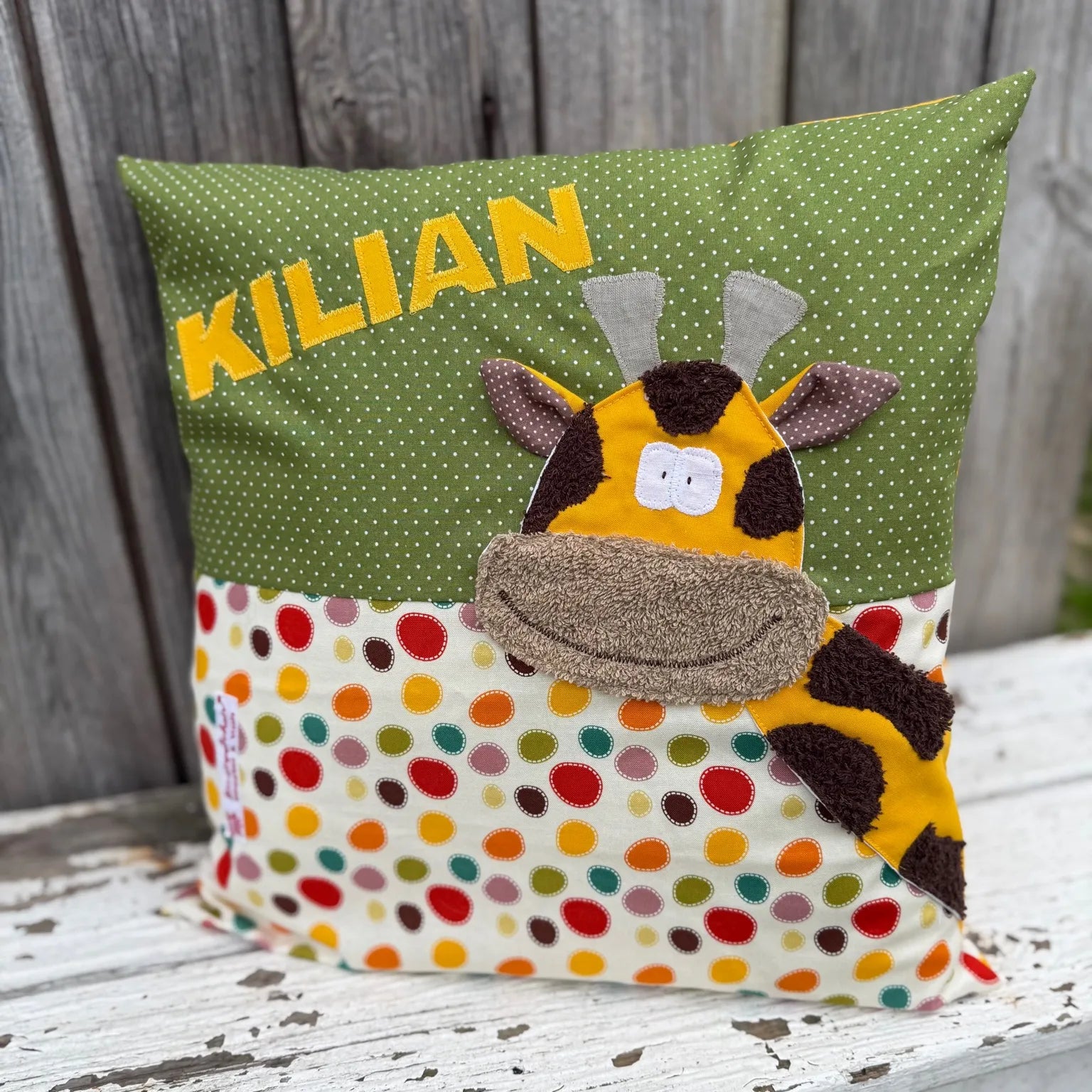 Handgenähtes Kinderkissen personalisiert, gestaltet grün mit weißen Punkten im oberen Teil, cremefarben mit bunten Tupfen im unteren Teil. Auf dem Kissen ist eine Giraffen-Applikation in leuchtendem Gelb. Fotografiert vor einem rustikalen Holzhintergrund 