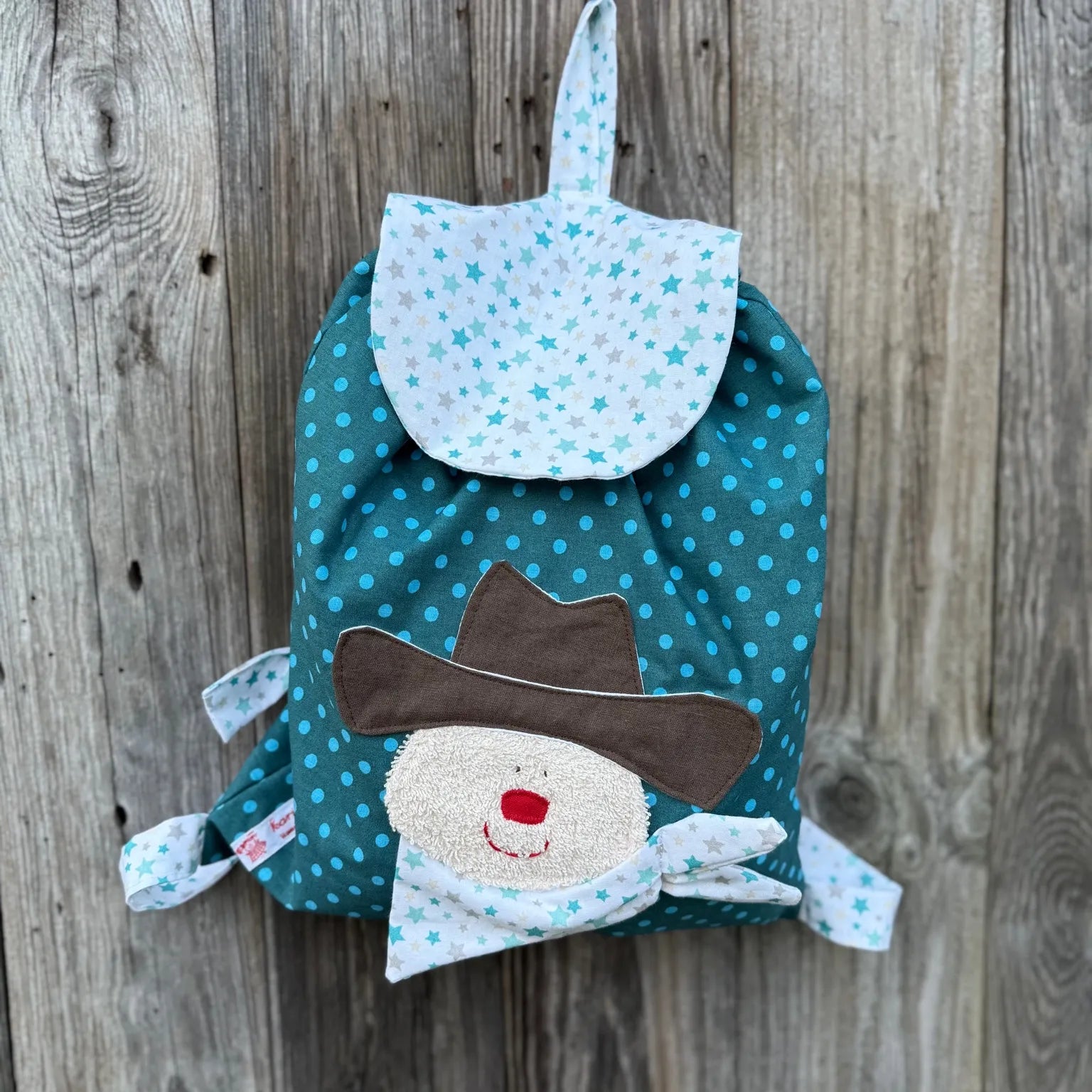 Handgemachter Kinderrucksack ist  petrol 
mit Punkten , hat eine weiße Klappe mit Sternen und eine aufgenähte Applikation eines Cowboys.
