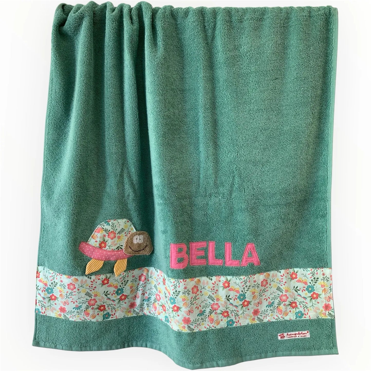 Handgemachtes mintfarbenes Duschtuch mit applizierter Schildkröte in der linken unteren Ecke, personalisiert mit Namen in rosa Buchstaben. Unten am Handtuch ist ein Streifen in bunt geblümt aufgenäht.