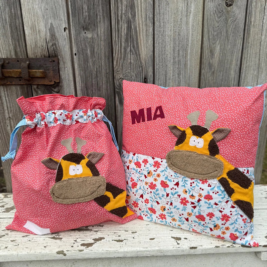 Handgemachtes Set bestehend aus einer Wetbag und einem personalisierten Kissen. Beide Artikel sind aus rosa Stoff mit weißen Punkten sowie floralen Mustern in Rot, Blau und Weiß gefertigt. Die Applikationen zeigen eine fröhliche Giraffe mit gelbem Gesicht, braunen Flecken und großem, beigem Maul. Das Kissen ist mit dem Namen „MIA“ in dunkelpinken Buchstaben personalisiert. Die Wetbag hat einen Tunnelzug mit blau-weiß gestreiften Bändern. Die Artikel stehen auf einer weiß gestrichenen, rustikalen Holzbank