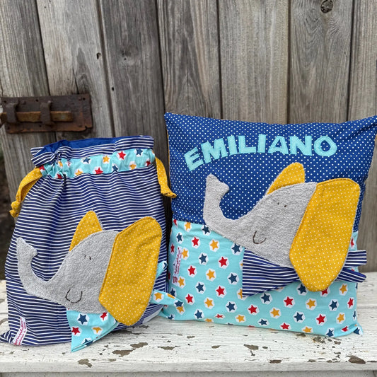 Handgemachtes Set bestehend aus einer Wetbag und einem personalisierten Kissen. Beide Teile sind aus blau-weiß gestreiftem Stoff und hellblauem Stoff mit bunten Sternen gefertigt. Auf beiden befindet sich eine freundliche Elefanten-Applikation mit grauem Gesicht, großen gelben Ohren und einem gemusterten Halstuch. Das Kissen ist zusätzlich mit dem Namen „EMILIANO“ in hellblauen Buchstaben personalisiert. Die Artikel stehen auf einer weiß gestrichenen, verwitterten Holzbank vor einer rustikalen Holzwan