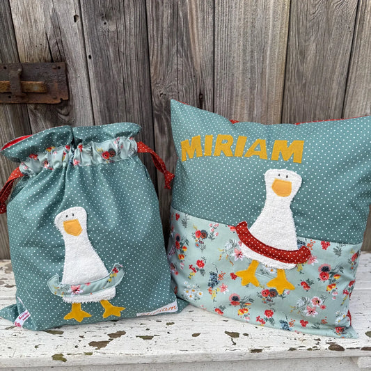 Handgefertigtes Set bestehend aus einer Wetbag und einem personalisierten Kissen. Beide Artikel sind aus mintgrünem Stoff mit weißen Punkten und floralen Mustern gefertigt. Auf beiden ist eine niedliche Gänse-Applikation mit orangefarbenem Schnabel, gelben Füßen und einem dekorativen Schwimmreifen zu sehen. Das Kissen ist mit dem Namen „MIRIAM“ in gelben Buchstaben personalisiert. Die Wetbag hat ein rotes Zugband mit weißen Punkten. Die Artikel stehen auf einer weiß gestrichenen, verwitterten Holzbank