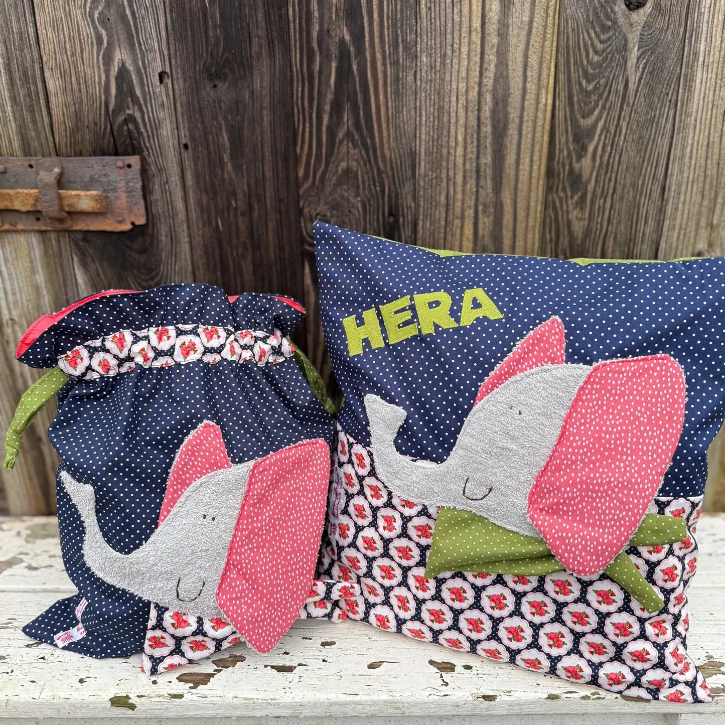  Handgemachtes Set aus Wetbag und personalisiertem Kissen, beide aus dunkelblauem Stoff mit weißen Punkten und floral gemustertem Stoff in Rot, Weiß und Schwarz. Beide Stücke zeigen eine Applikation eines fröhlichen Elefanten mit grauem Körper, pink gepunkteten Ohren und grünem Halstuch. Das Kissen ist mit dem Namen „HERA“ in grünen Buchstaben personalisiert. Die Artikel stehen auf einer weißen, verwitterten Holzbank vor einer rustikalen Holzwand.