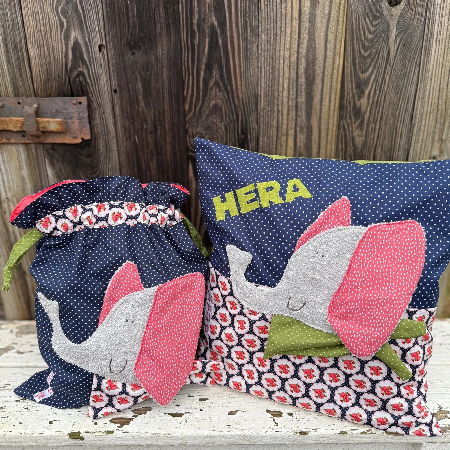  Handgemachtes Set aus Wetbag und personalisiertem Kissen, beide aus dunkelblauem Stoff mit weißen Punkten und floral gemustertem Stoff in Rot, Weiß und Schwarz. Beide Stücke zeigen eine Applikation eines fröhlichen Elefanten mit grauem Körper, pink gepunkteten Ohren und grünem Halstuch. Das Kissen ist mit dem Namen „HERA“ in grünen Buchstaben personalisiert. Die Artikel stehen auf einer weißen, verwitterten Holzbank vor einer rustikalen Holzwand.