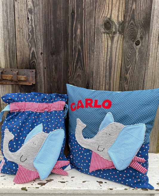 Handgemachtes Set aus einer Wetbag und einem personalisierten Kissen. Beide bestehen aus blauem Stoff mit kleinen Herzmustern und weißen Punkten. Die Applikationen zeigen einen freundlichen grauen Elefanten mit großen, hellblau-weiß gestreiften Ohren und einem roten Halstuch mit Karomuster. Das Kissen ist zusätzlich mit dem Namen „CARLO“ in roten Buchstaben personalisiert. Die Artikel stehen auf einer weiß gestrichenen, rustikalen Holzbank vor einer alten Holzwand.