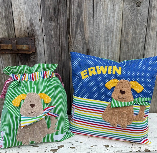  Handgefertigtes Set bestehend aus einer Wetbag und einem personalisierten Kissen. Die Wetbag ist aus grün-weiß gestreiftem Stoff mit buntem Streifenmuster am oberen Rand gefertigt. Das Kissen kombiniert blauen Punktestoff mit einem bunten Streifenmuster. Beide Teile zeigen eine Applikation eines freundlichen braunen Hundes mit gelben Ohren und Halstuch. Das Kissen ist mit dem Namen „ERWIN“ in gelben Buchstaben personalisiert. Die Artikel stehen auf einer weiß gestrichenen, verwitterten Holzbank vor 
