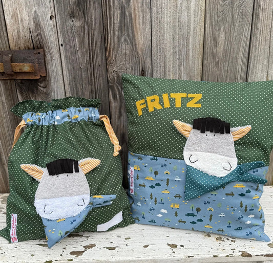  Handgefertigtes Set bestehend aus einer Wetbag und einem personalisierten Kissen. Beide Teile bestehen aus grünem Stoff mit weißen Punkten und hellblauem Stoff mit Fahrzeug- und Naturmotiven. Auf beiden ist eine niedliche Esel-Applikation mit grauem Gesicht, schwarzer Mähne, gestreiften Ohren und Halstuch angebracht. Das Kissen ist mit dem Namen „FRITZ“ in gelben Buchstaben personalisiert. Die Artikel stehen auf einer weiß gestrichenen, rustikalen Holzbank vor einer alten Holzwand.