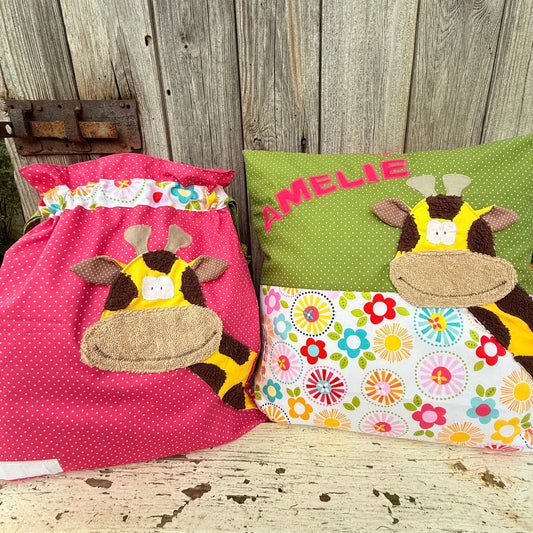 Handgefertigtes Set bestehend aus einer Wetbag und einem personalisierten Kissen. Die Wetbag ist aus pinkem Stoff mit weißen Punkten gefertigt und hat einen bunten Stoffstreifen mit Blumenmuster am oberen Rand. Beide Teile zeigen eine fröhliche Giraffen-Applikation mit gelbem Körper, braunen Flecken und beigem Maul. Das Kissen besteht aus grünem Punktestoff und einem unteren Bereich mit buntem Blumenmuster und ist mit dem Namen „AMELIE“ in pinken Buchstaben personalisiert. Die Artikel stehen auf einer