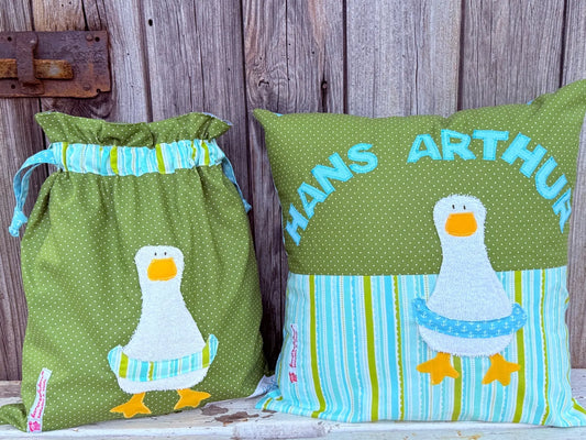 Handgemachtes Set bestehend aus einer Wetbag und einem personalisierten Kissen. Beide Artikel sind aus grünem Stoff mit weißen Punkten und blau-grün gemustertem Stoff gefertigt. Auf beiden ist eine fröhliche Gänse-Applikation mit orangefarbenem Schnabel und Füßen sowie einem gestreiften Schwimmreifen zu sehen. Das Kissen trägt den Namen „HANS ARTHUR“ in großen, türkisfarbenen Buchstaben. Die Artikel stehen auf einer weiß gestrichenen, verwitterten Holzbank vor einer rustikalen Holzwand.