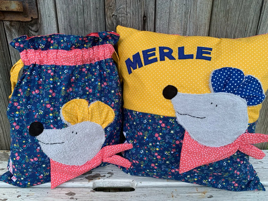  Handgemachtes Set bestehend aus einer Wetbag und einem personalisierten Kissen. Beide Teile sind aus dunkelblauem Stoff mit buntem Blumenmuster sowie gelbem und pinkem Punktestoff gefertigt. Auf beiden ist eine fröhliche Maus-Applikation mit grauem Gesicht, farbigen Ohren (gelb oder blau) und rosa Halstuch zu sehen. Das Kissen trägt den Namen „MERLE“ in blauen Buchstaben. Die Artikel stehen auf einer weiß gestrichenen, verwitterten Holzbank vor einer rustikalen Holzwand.