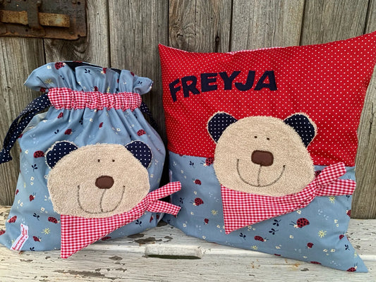 Handgefertigtes Set bestehend aus einer Wetbag und einem personalisierten Kissen. Beide Artikel sind aus hellblauem Stoff mit Marienkäfer-Muster sowie rotem und dunkelblauem Stoff mit Punkten gefertigt. Auf beiden ist eine freundliche Bären-Applikation mit beigefarbenem Gesicht, dunkelblauen Ohren und einem roten Halstuch mit Karomuster angebracht. Das Kissen ist zusätzlich mit dem Namen „FREYJA“ in dunkelblauen Buchstaben personalisiert. Die Artikel stehen auf einer weiß gestrichenen, rustikalen Holz