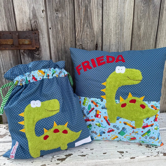  Handgefertigtes Set bestehend aus einer Wetbag und einem personalisierten Kissen. Beide Teile sind aus blauem Stoff mit weißen Punkten sowie hellblauem Stoff mit buntem Fahrzeugmuster gefertigt. Die Applikationen zeigen einen fröhlichen grünen Dinosaurier mit weißen Augen, gelben Zacken und roten Punkten auf dem Rücken. Das Kissen ist mit dem Namen „FRIEDA“ in roten Buchstaben personalisiert. Die Wetbag hat ein grünes Zugband mit weißen Streifen. 